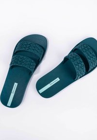Infradito in gomma turchese con cinturini intrecciati, soletta decorata e un accento blu chiaro del logo sul lato, caratterizzati da un design piatto e casual.