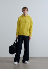 Κίτρινο φούτερ με κορδόνια, εμπρόσθια τσέπη; μαύρο παντελόνι με φαρδιά μπατζάκια; κρατάει μια μαύρη σακούλα duffel; λευκά αθλητικά παπούτσια; λείες υφές, casual στυλ.