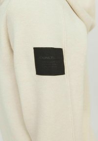 Manche en maille beige avec une étiquette en tissu noire rectangulaire cousue près de la partie supérieure du bras.