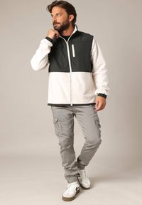 Homme barbu portant une veste polaire zippée noire et blanche, un pantalon cargo gris et des baskets blanches, posant avec une main près de son visage.