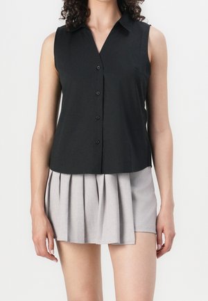 Person wearing a sleeveless black button-up top and a light grey pleated mini skirt, standing against a plain background.