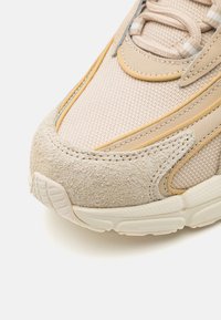 Beige athletic shoe featuring a mesh upper with suede accents, rounded toe, textured sole, and stitched detailing in yellow and gray.