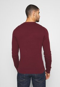 Suéter de punto burdeos con un sencillo cuello redondo y puños de canalé, combinado con unos jeans de mezclilla, visto desde atrás.