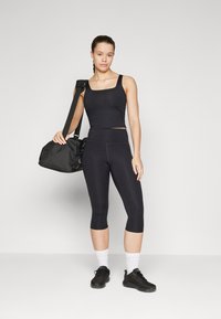 Svart sportbh med fyrkantig halsringning, tillsammans med svarta cropped leggings. Modellen bär svarta sneakers och håller en svart duffelväska.