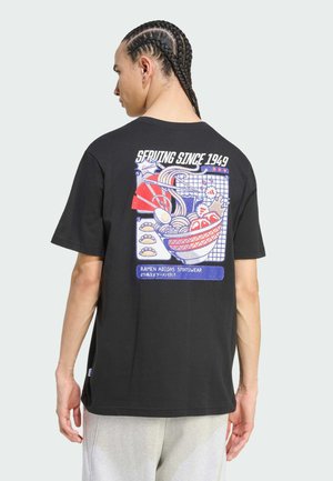 Person med flettet hår iført en sort T-shirt med en farverig grafik af ramen og sushi med teksten "Serving Since 1949" på ryggen.