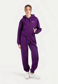 Sweat à capuche violet zippé avec poches avant et pantalon de survêtement assorti. Le matériau semble doux, avec des poignets élastiques et des détails de logo brodés subtils.