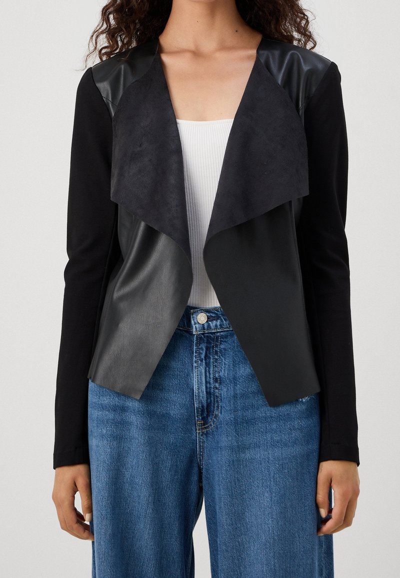 Femme portant une veste noire ouverte sur l'avant avec des matériaux mélangés sur un top blanc côtelé et un jean bleu taille haute, montrée des épaules aux hanches.