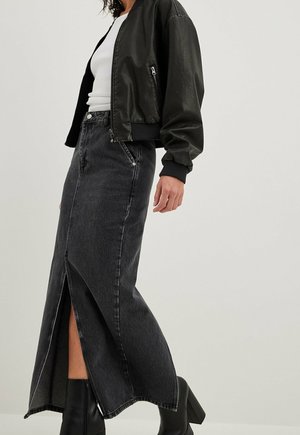Femme portant une veste en cuir noire, un haut blanc, une longue jupe en jean noire avec une fente à l’avant, et des bottines noires à talons sur un fond uni.