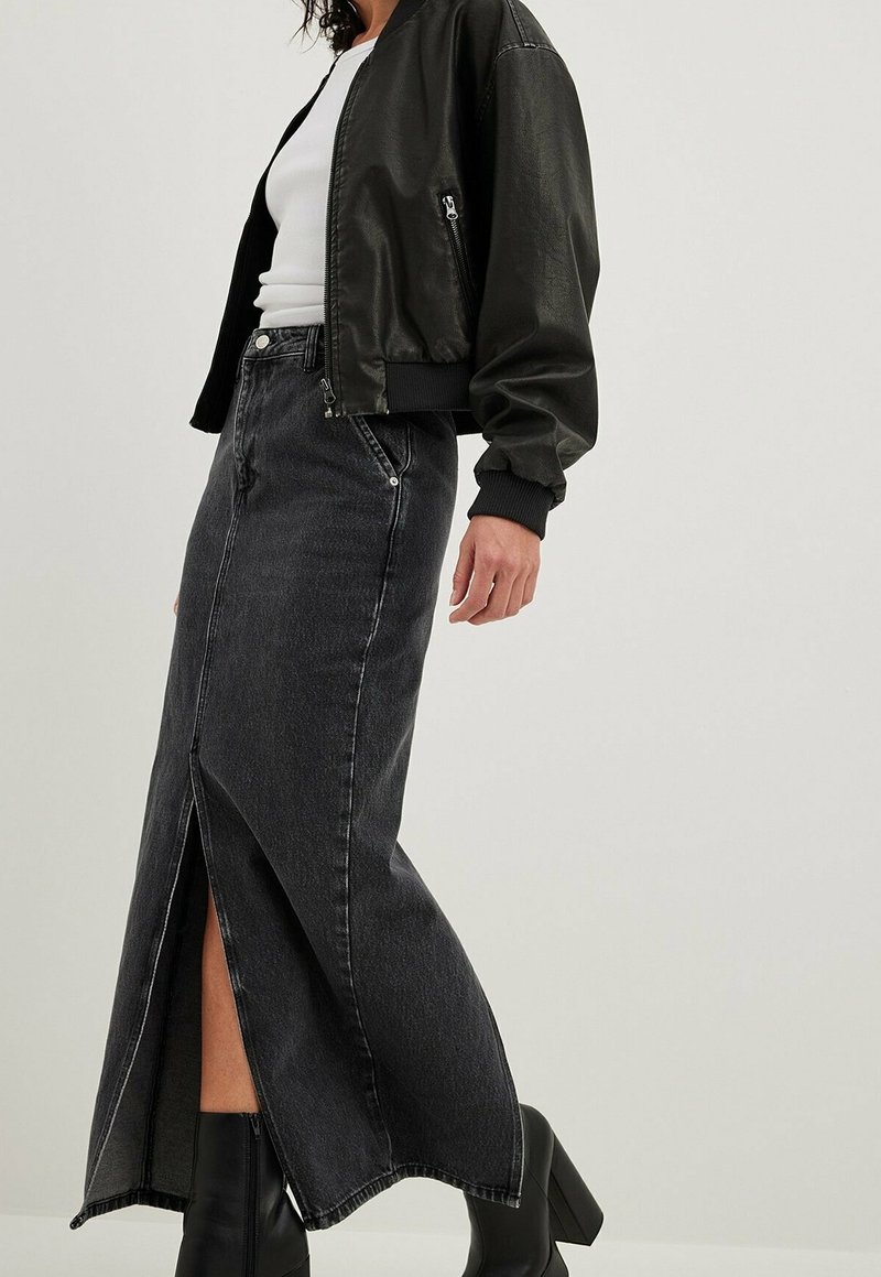 Femme portant une veste en cuir noire, un haut blanc, une longue jupe en jean noire avec une fente à l’avant, et des bottines noires à talons sur un fond uni.