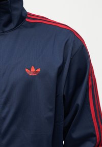 Marineblå jakke med røde striper på ermene og en rød trefoil-logo på venstre bryst, med høy krage.