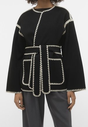 Chaqueta negra con cinturón y ribete en zigzag blanco en las costuras, puños, bolsillos y bordes, usada sobre pantalones oscuros en una persona de pie.