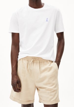 Homme portant un t-shirt blanc à manches courtes avec un petit motif bleu et un short beige à cordon de serrage avec ourlets retroussés.