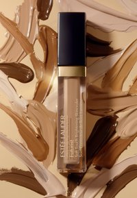 Estee Lauder Futurist concealer i en klar tube med svart hette og gulldetaljer, plassert på en bakgrunn av sammenblandet beige og brune toner.