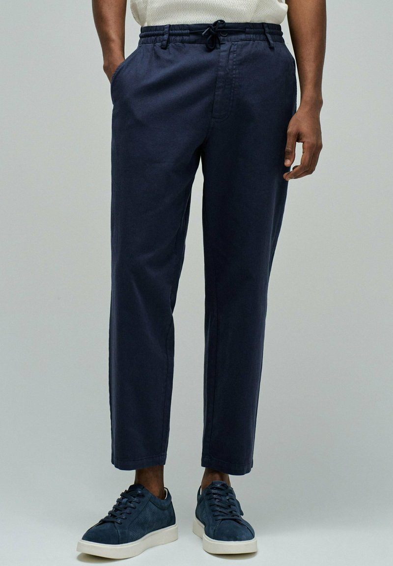 Hombre vestido con pantalones azul marino con cordón y zapatillas azul marino con suelas blancas, de pie contra un fondo gris liso.