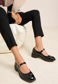 Črne Mary Jane čevlje iz lakiranega usnja z okroglim prstom, trakcem okoli gležnja s srebrno sponko in nizkim debelim petnim delom. Gladek tekstura.