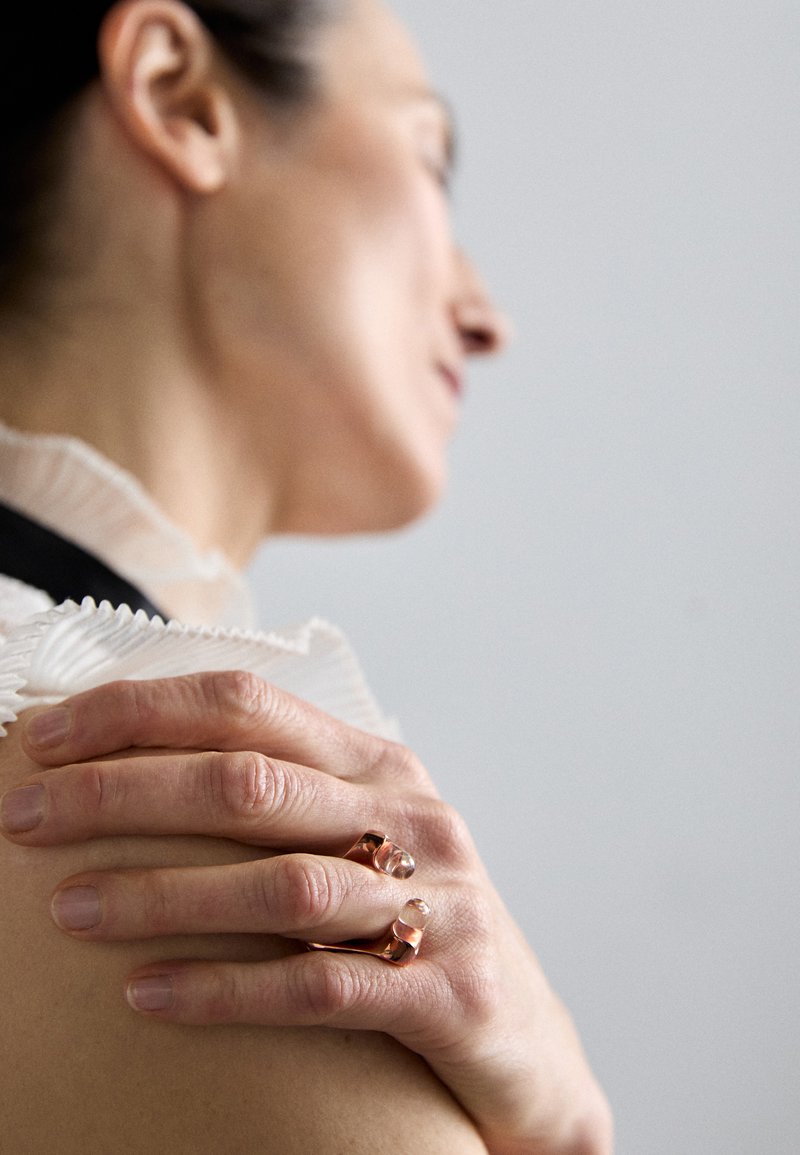 Anelli in oro rosato su dita, con un design liscio e curvo e accenti in pietra trasparente. Una delicata trama di tessuto è visibile sullo sfondo.