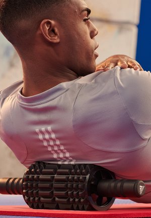 Gray textured athletic shirt with white stripes, paired with a black foam roller featuring a patterned surface and ergonomic grips, on a red mat.