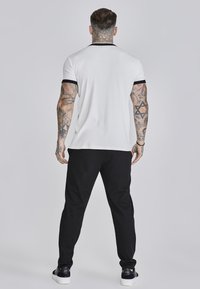 Witte polo met zwarte accenten, gecombineerd met zwarte trainingsbroeken en zwarte sneakers. Heeft een getailleerd ontwerp waarbij getatoeëerde armen zichtbaar zijn.