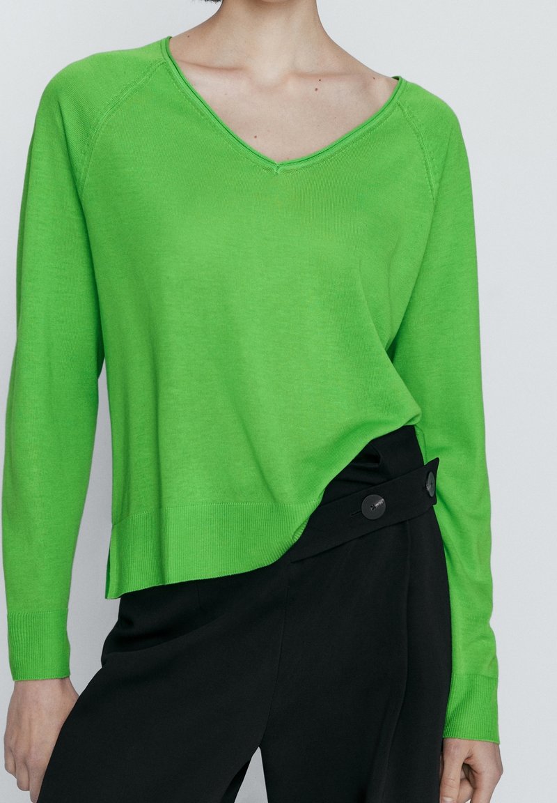Femme portant un pull vert vif à col en V, rentré dans un pantalon noir taille haute avec une large ceinture boutonnée.