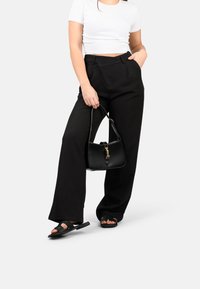 Pantaloni neri a gamba larga con un taglio sartoriale e tasche frontali, abbinati a una borsa nera con dettagli dorati. Il modello indossa sandali neri.