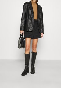 Schwarze Lederblazer, brauner Rollkragenpullover, schwarzer Minirock und knielange schwarze Stiefel. Accessoires umfassen eine schwarze Tasche mit strukturiertem Finish.