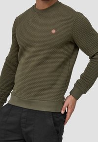 Oliwkowy sweter z fakturowanej tkaniny, okrągłym dekoltem, ściągaczami przy mankietach i w pasie. Posiada małą skórzaną naszywkę z logo na klatce piersiowej.