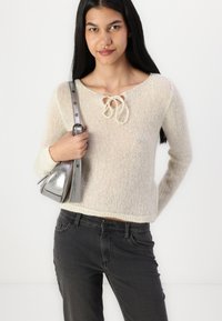 Lyse beige strikket sweater med rund hals og bindedetalje, parret med mørke grå jeans og en metallisk sølvhåndtaske.