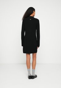 Μαύρο πλεκτό φόρεμα με ριμπ ύφανση, μακριά μανίκια και μήκος μέχρι το μέσο του μηρού. Διαθέτει μικρό λογότυπο στο πίσω μέρος και φοριέται με γκρι κάλτσες και μαύρα παπούτσια.