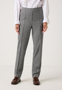 Vrouw in hoge, grijze geruite broek met voorplooien, witte blouse met gerimpelde manchetten en bruine loafers staand tegen een effen achtergrond.
