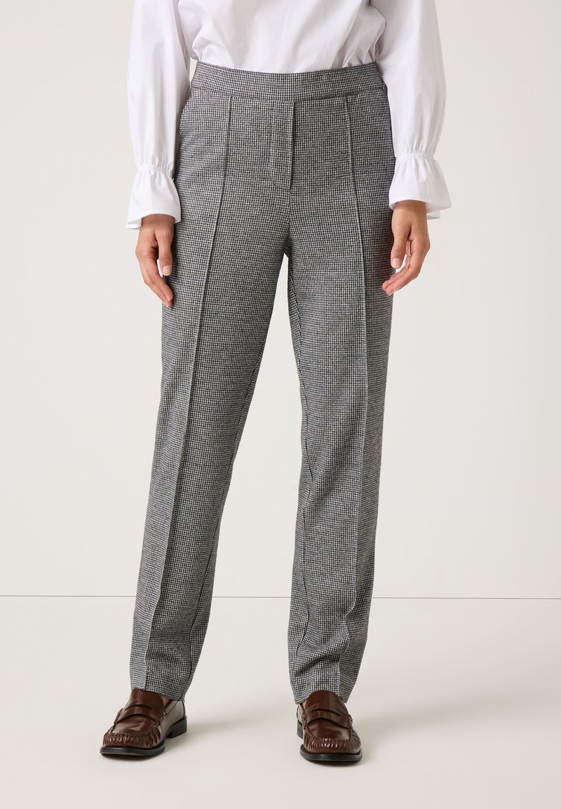 Vrouw in hoge, grijze geruite broek met voorplooien, witte blouse met gerimpelde manchetten en bruine loafers staand tegen een effen achtergrond.