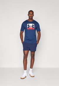 Marineblå t-shirt med stort Under Armour-logo, parret med marineblå shorts. Modellen bærer hvide sneakers med tan såler og hvide sokker.