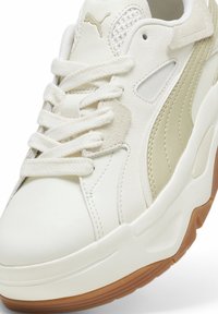 Baskets Puma blanches et beiges avec semelle épaisse en gomme, détails en mesh et daim, et lacets blancs noués en nœud à l'avant.