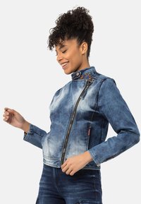 Jeansjacke mit einem ausgewaschenen blauen Farbton, hohem Kragen, asymmetrischem Frontreißverschluss und gemustertem Besatz an der Schulter. Lange Ärmel und schmaler Schnitt.