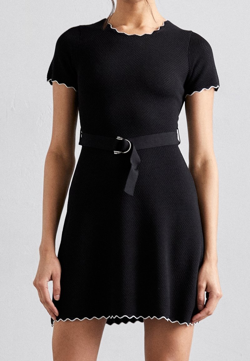 Femme portant une robe noire texturée à manches courtes avec des bords festonnés blancs, un col rond et une ceinture noire avec une boucle en anneau métallique.