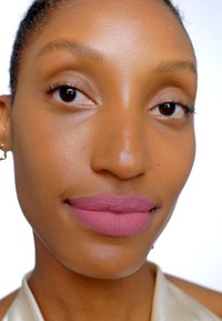 Närbild av ett ansikte med slät hy, där fulla läppar är målade med dämpad rosa matt läppstift och naturlig ögonmakeup mot en ljus bakgrund.