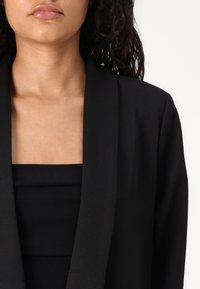 Černý otevřený blazer s hladkým povrchem, s širokým límcem a přiléhavým černým topem pod ním.