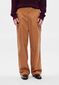 Barna pamut nadrág, laza szabással, elöl redőkkel és gombos rögzítéssel. Sötétbarna cipővel viselve.