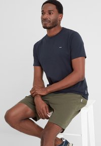 Navy short-sleeve t-shirt with a small white logo, paired with olive green shorts. Model seated on a white stool against a light background.
