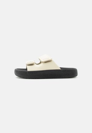 Beige slidssandal med to justerbare velcrolukninger og tyk sort sål med tekstureret mønster, isoleret på hvid baggrund.