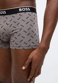 Gros plan sur une personne portant un boxer gris orné de logos noirs répétitifs "BOSS" et d'une ceinture noire avec l'inscription "BOSS".