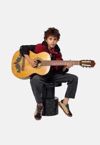 Kind dat een rode vest over een zwarte jas draagt, met een houten akoestische gitaar met ingewikkelde zwarte ontwerpen, zittend op een zwart gespikkelde kruk.