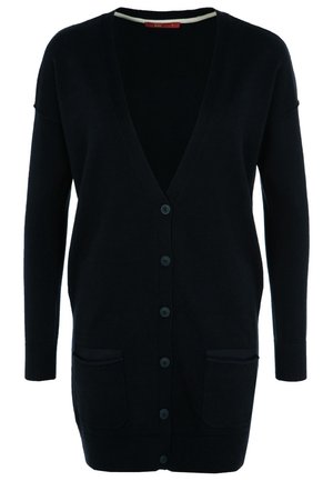 Femme portant un long cardigan noir boutonné avec des poches avant, sur un haut blanc et un jean bleu, debout devant un fond uni.
