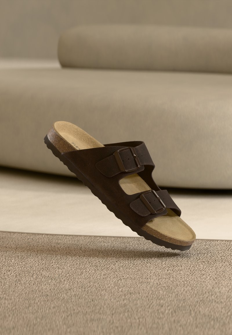 Sandale mule en daim marron avec deux boucles ajustables, semelle intérieure profilée et semelle extérieure en caoutchouc texturé, sur fond neutre.