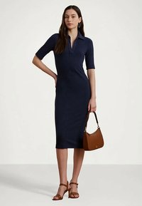 Kvinna i en marinblå ribbad midiklänning, bruna klacksandaler och matchande brun handväska, stående med en hand på höften.