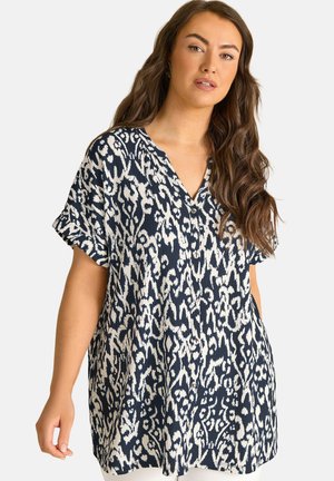 Femme portant un chemisier ample à manches courtes avec un imprimé animal abstrait noir et blanc, posant devant un fond clair uni.