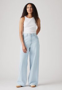 Light blue wide-leg jeans in denim, high-waisted design with a button closure and front pockets; model wearing a white sleeveless top.