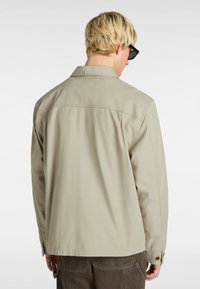 Light beige long-sleeve jacket with a collar, back yoke seam, and buttoned cuffs. Smooth, woven fabric with subtle texture.