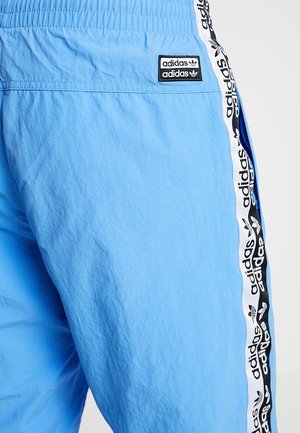 Jasnoniebieskie spodnie dresowe Adidas z elastycznym pasem, z białym i czarnym markowanym paskiem po boku oraz małą naszywką z logo Adidas przy kieszeni.