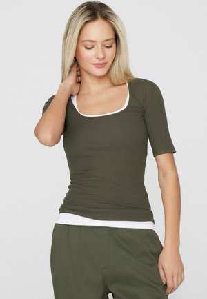 Mladá žena s dlouhými blond vlasy, která má na sobě olivově zelené přiléhavé tričko s krátkým rukávem přes bílé tílko a odpovídající olivové kalhoty.