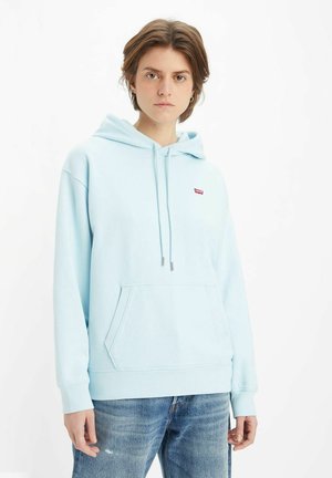 Felpa blu chiaro con tasca a marsupio, cappuccio con cordino e piccolo logo rosso sul petto. Tessuto morbido e vestibilità comoda. Indossata con jeans strappati.